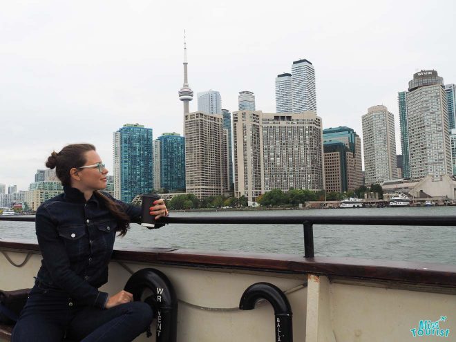 Our collegue, Yulia, sitting on a boat, holding a cup, with the Toronto skyline in the background, highlighting the CN Tower and waterfront skyscrapers
