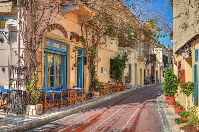 A quaint street in Athens with colorful buildings and empty wooden chairs lining the sidewalk, exuding a serene Mediterranean atmosphere
