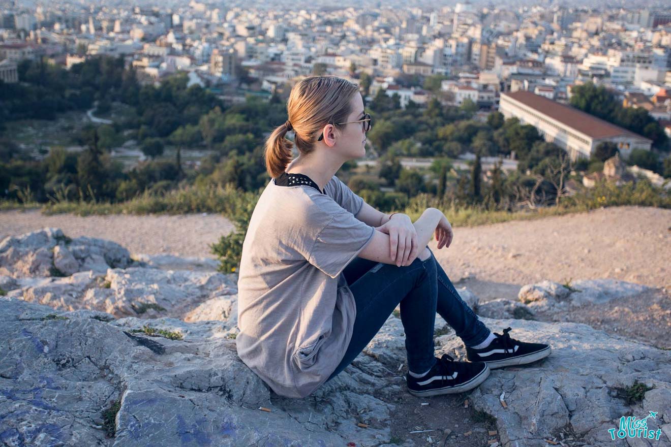 The writer of the post sitting on a rocky outcrop overlooking the sprawling city of Athens during sunset, reflecting a moment of quiet contemplation amidst urban exploration.