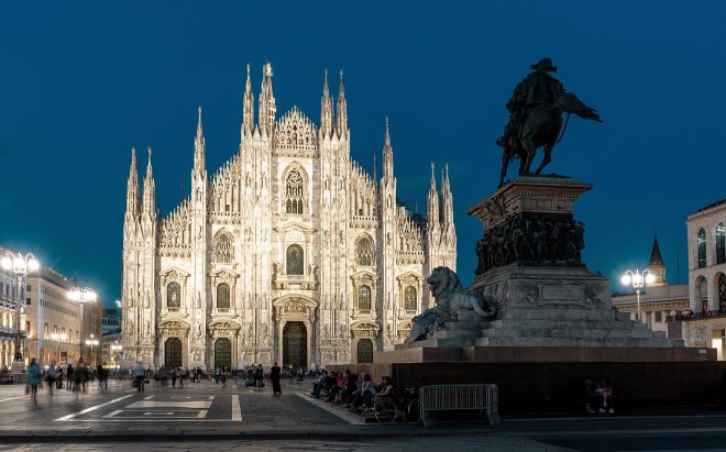 The Milan Cathedral (Duomo di Milano) stands illuminated at twilight, showcasing its intricate Gothic architecture and spires, with the statue of King Victor Emmanuel II dominating the foreground against a lively piazza scene
