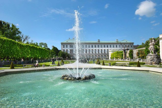 A classical fountain in the foreground with the Mirabell Palace and gardens in Salzburg under a clear sky