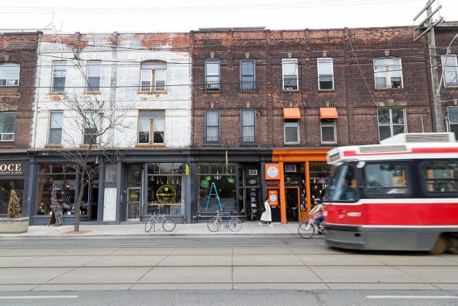 A daytime street view in Toronto showcasing a row of eclectic storefronts with a red and white bus passing by