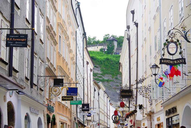 Cobbled street in Salzburg with traditional signage and the Hohensalzburg Fortress visible on the hill above