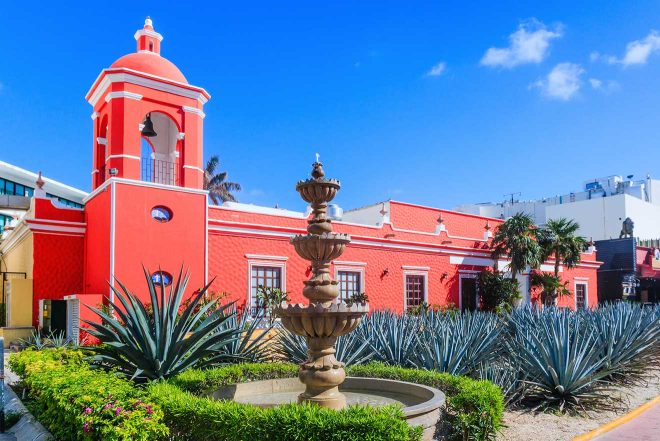 Vibrant red colonial-style building with a bell tower and fountain in the foreground, surrounded by agave plants, under a clear blue sky in Cancun, Mexico