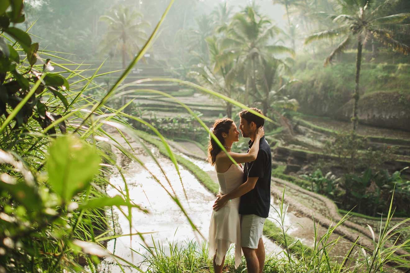 A couple stands closely together in an embrace, surrounded by lush greenery and terraced fields in the background.