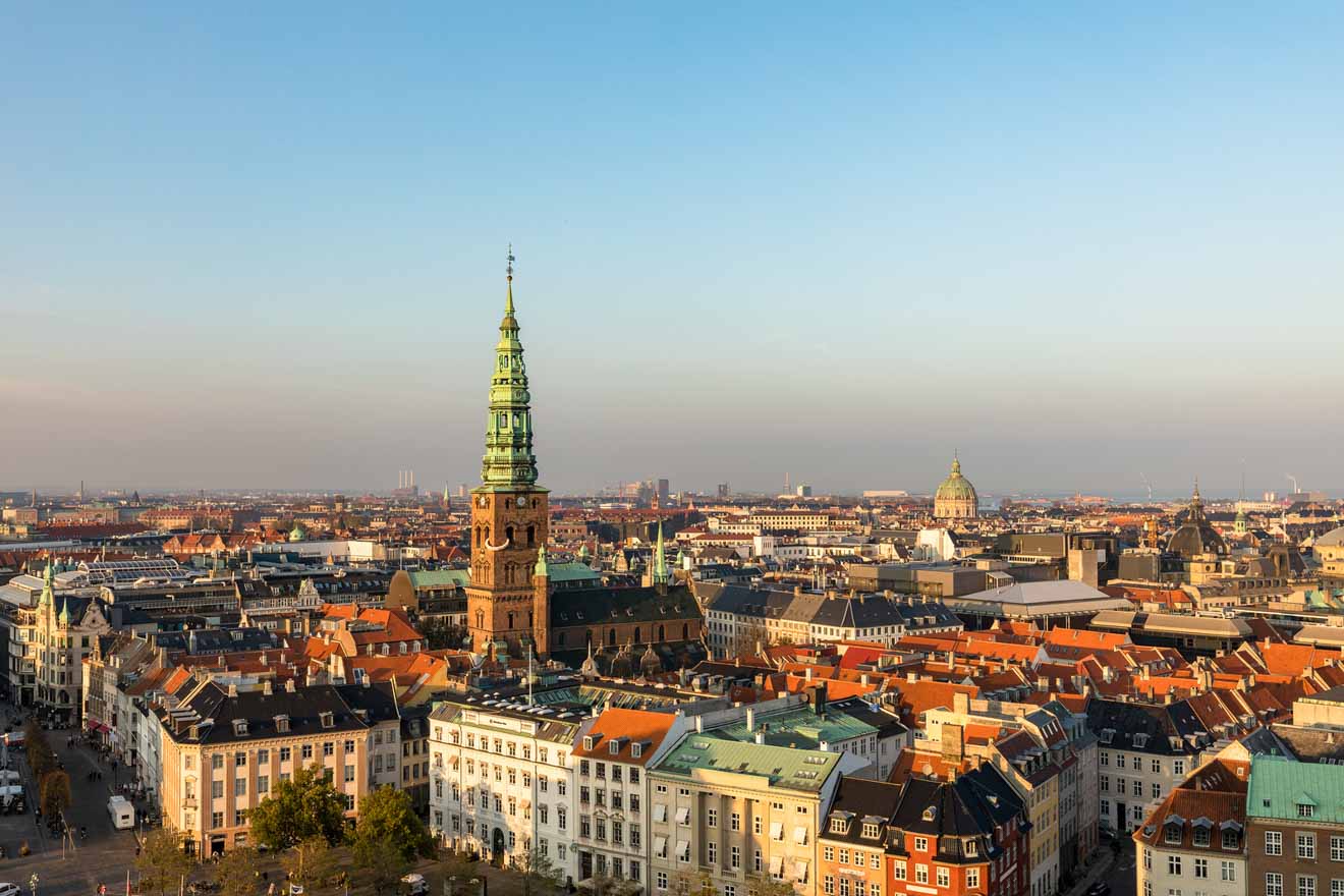Elevated view of central Copenhagen's rooftops and spires, bathed in the warm glow of a setting or rising sun, showcasing the city's blend of history and modernity.