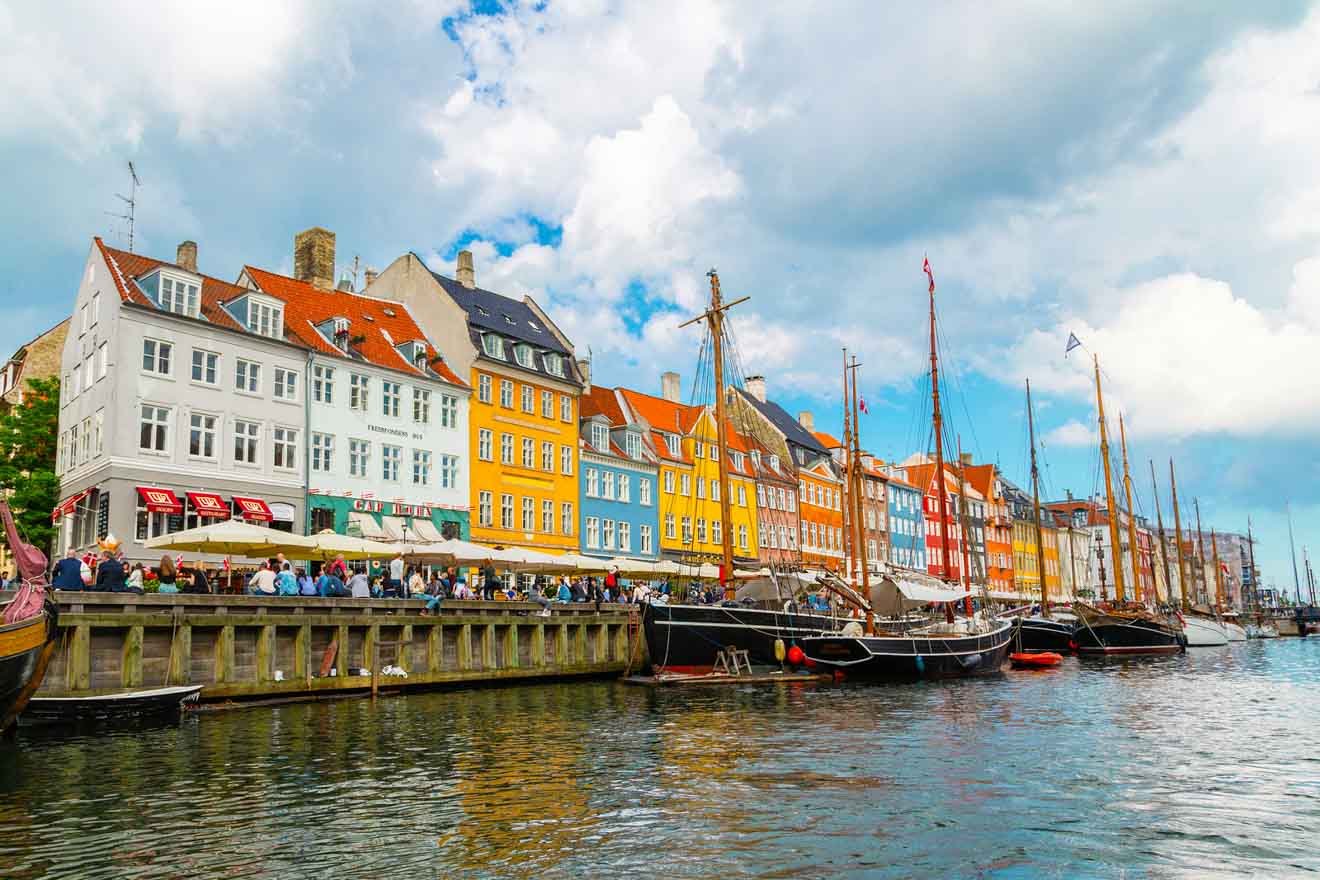 Colorful buildings line the waterfront in Nyhavn, Copenhagen, with anchored boats in the foreground and a partly cloudy sky overhead.