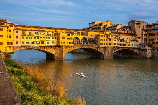 The famous Ponte Vecchio bridge spanning the Arno River in Florence, lined with shops and bustling with activity, with a single rowboat gliding underneath