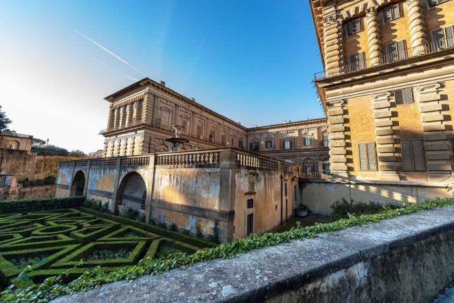 The imposing facade of Palazzo Pitti in Florence, viewed from its elaborate Renaissance garden, with geometric hedge patterns and a clear blue sky above.