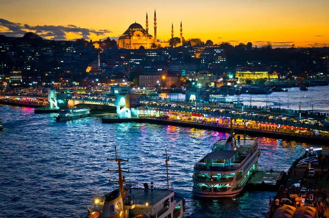 Nighttime view of Istanbul's waterfront with the Suleymaniye Mosque illuminated in the background, ferries docked at the port, and city lights reflecting off the Bosphorus