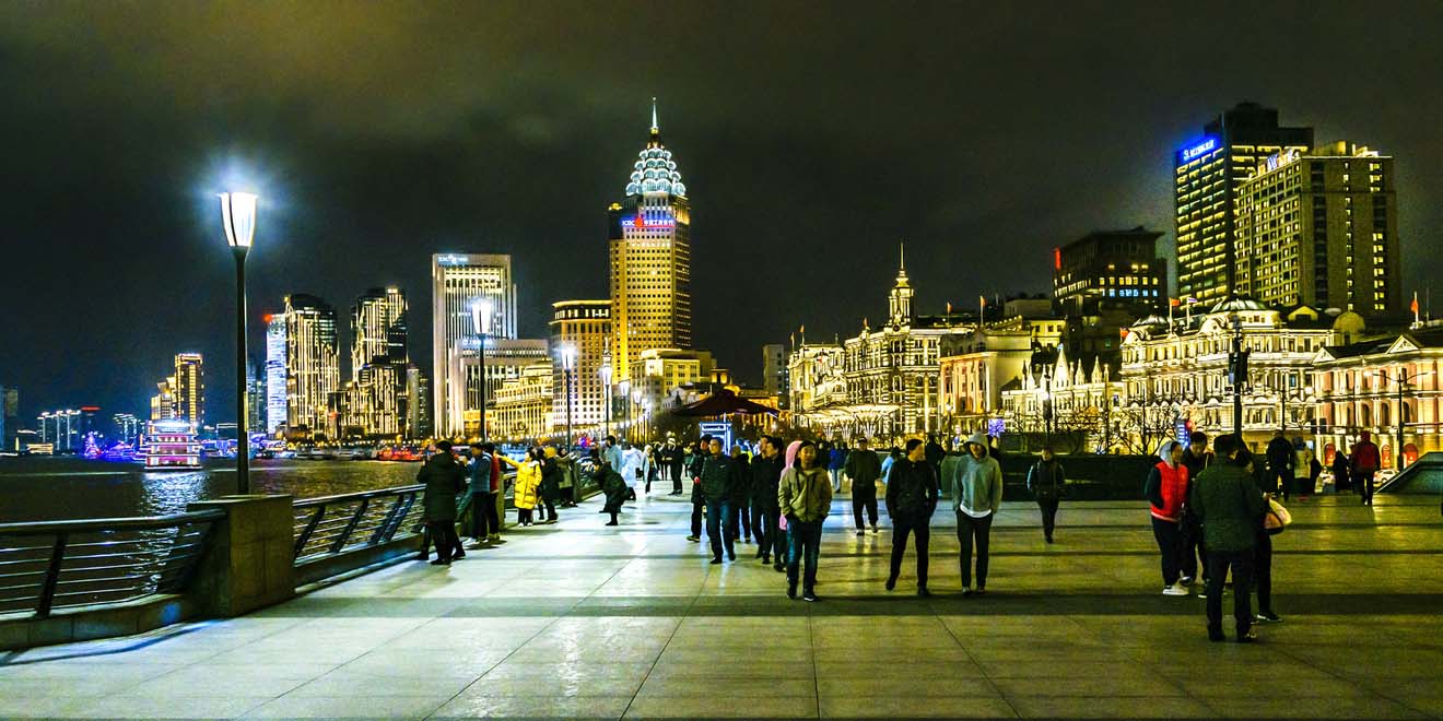 The bustling Bund waterfront promenade in Shanghai at night, with historic buildings lit up against the dark sky