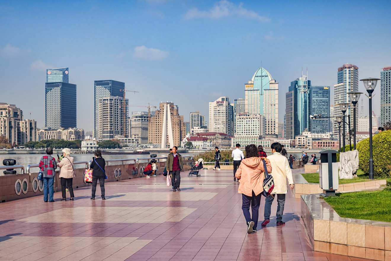 Crowds walking along the Shanghai Bund, with a view of the modern Pudong skyline across the Huangpu River during daytime.