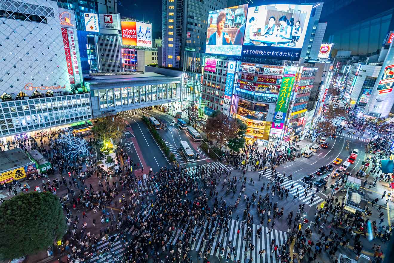 Aerial view of Shibuya Crossing at night, showcasing dense crowds of pedestrians and surrounded by illuminated skyscrapers