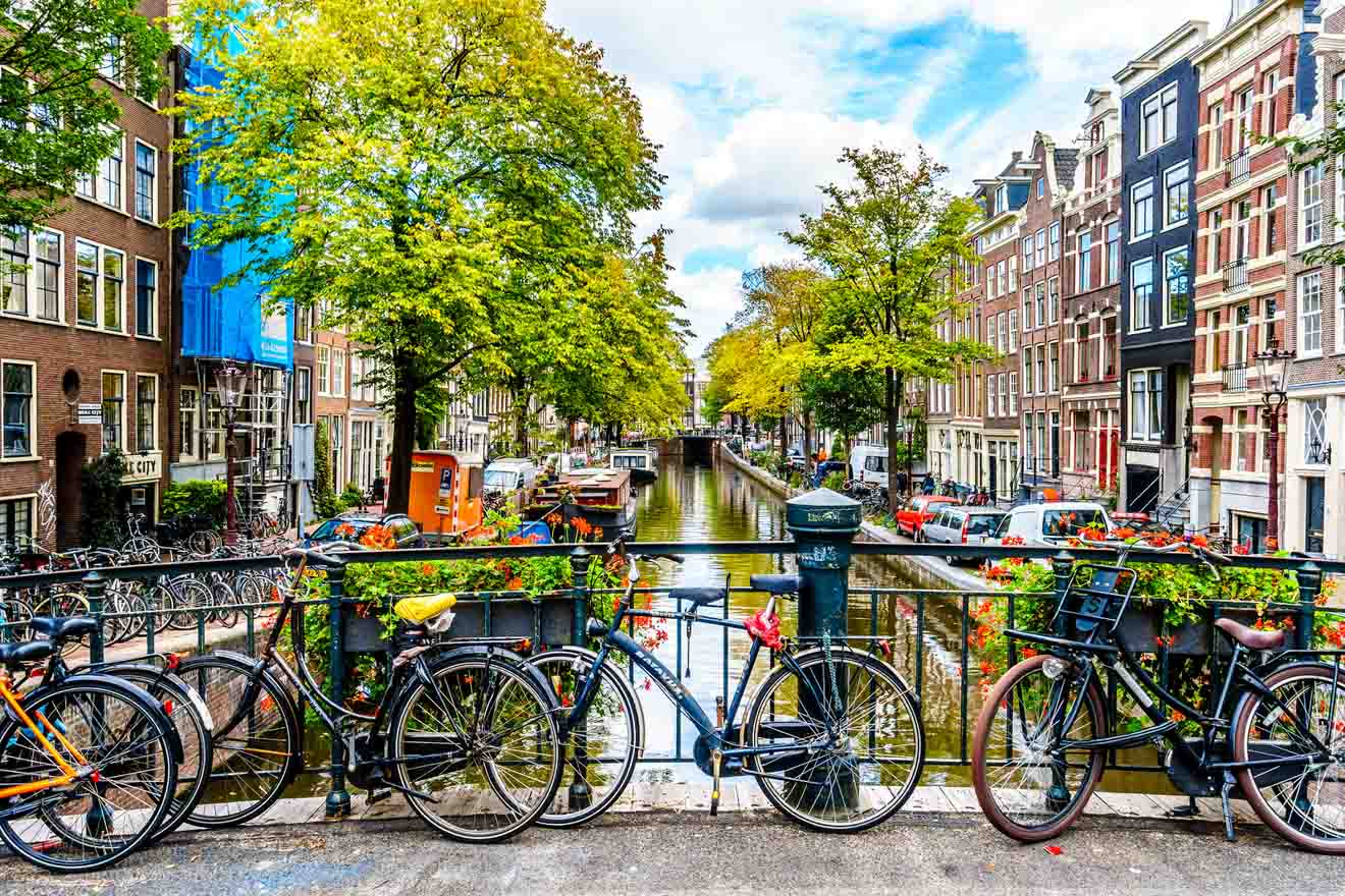 A picturesque Amsterdam canal lined with vibrant green trees and traditional Dutch architecture, with bicycles parked along a flower-adorned bridge railing in the foreground