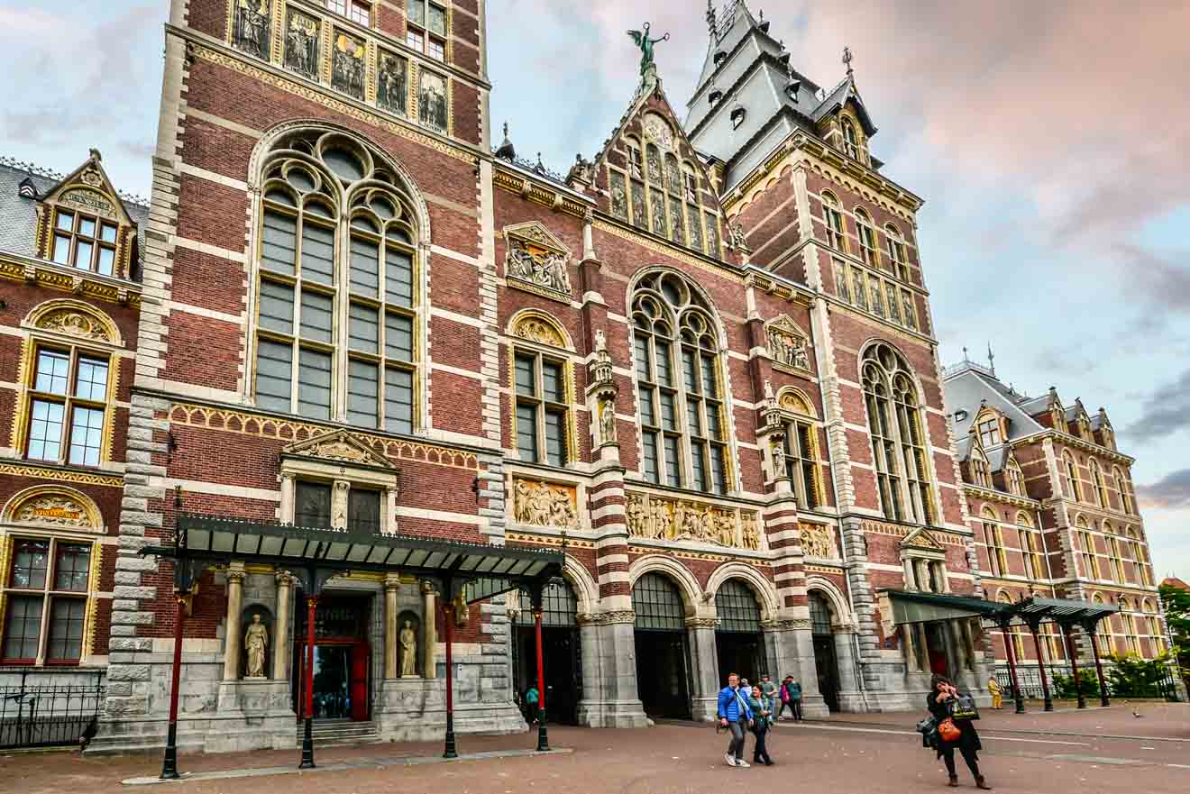 The Rijksmuseum's grand exterior in Amsterdam, showcasing its intricate Renaissance architecture with visitors milling about its entrance, under a sky hinting at the onset of dusk