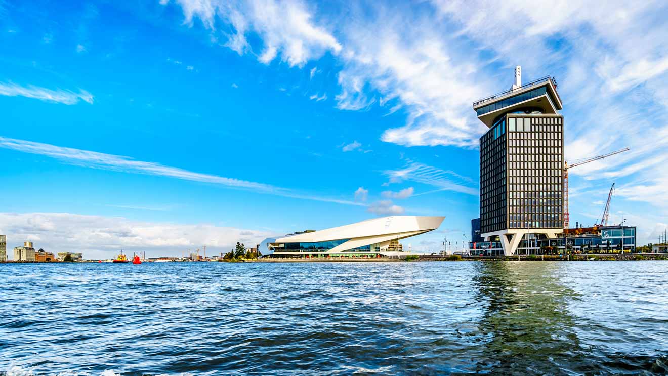 A panoramic view of Amsterdam's waterfront showcasing modern architecture with the EYE Film Institute resembling a white ship and the A'DAM Tower standing tall against a vibrant blue sky with scattered clouds