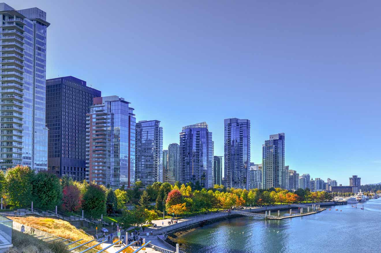 Clear blue sky over a waterfront pathway lined with autumn-colored trees and modern high-rise buildings in Vancouver.