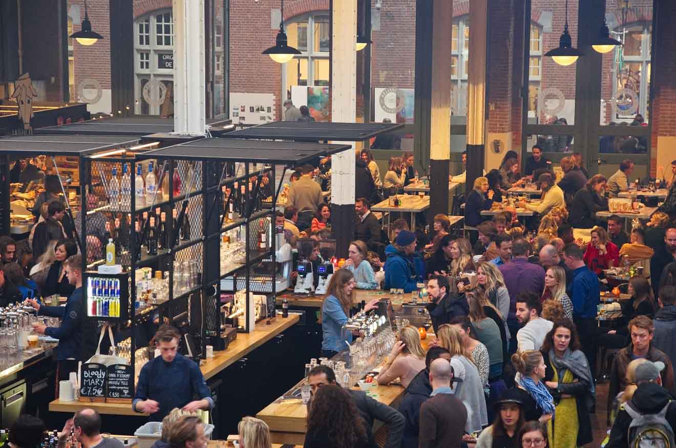 A bustling indoor food market atmosphere captured in Amsterdam, with patrons engaged in lively conversations around densely packed tables, and vendors busy at work in stalls offering a variety of culinary delights