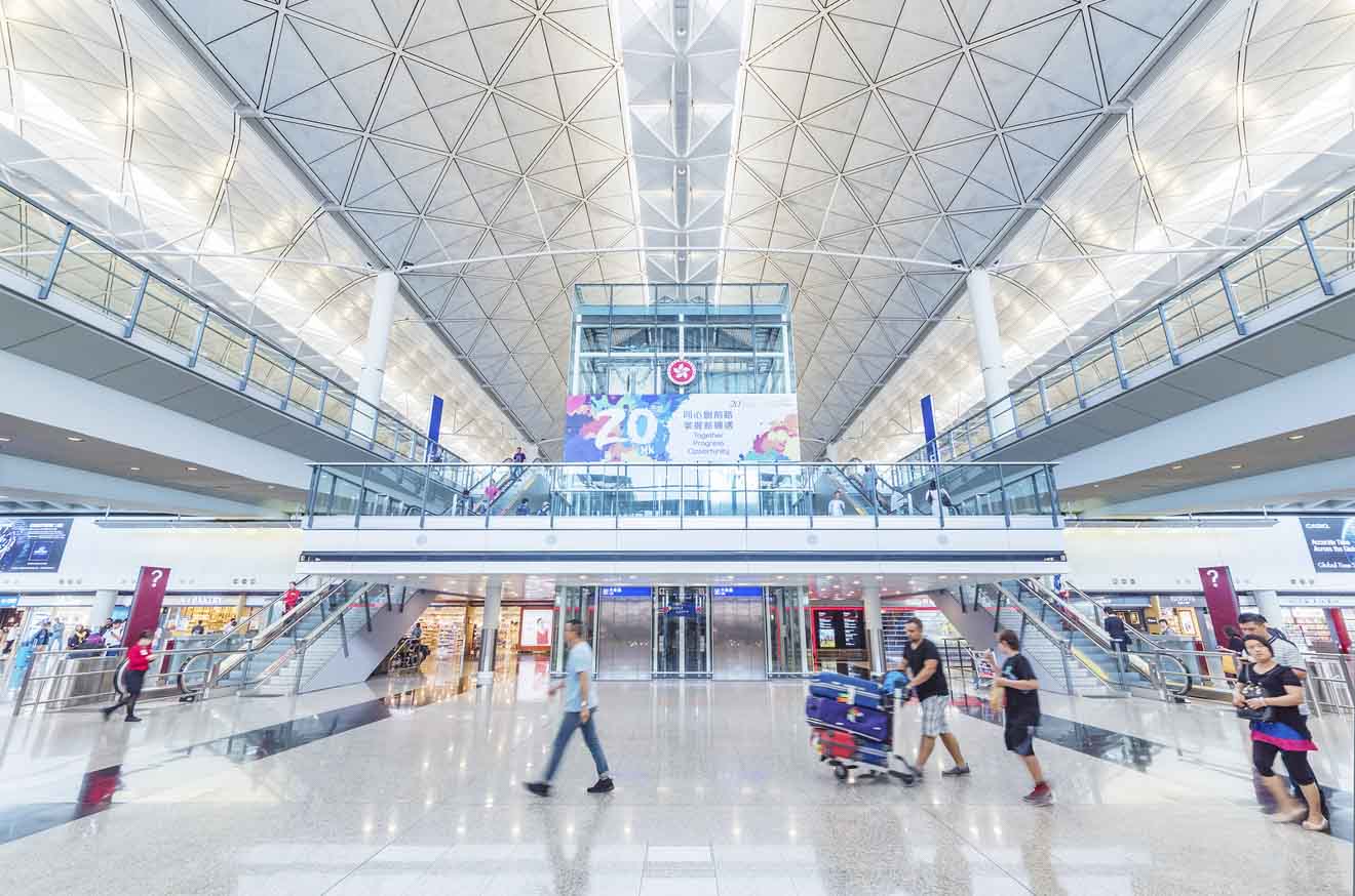 The spacious interior of Hong Kong's airport with passengers, a high ceiling, and the information board visible, reflecting the airport's modern design and busy atmosphere.