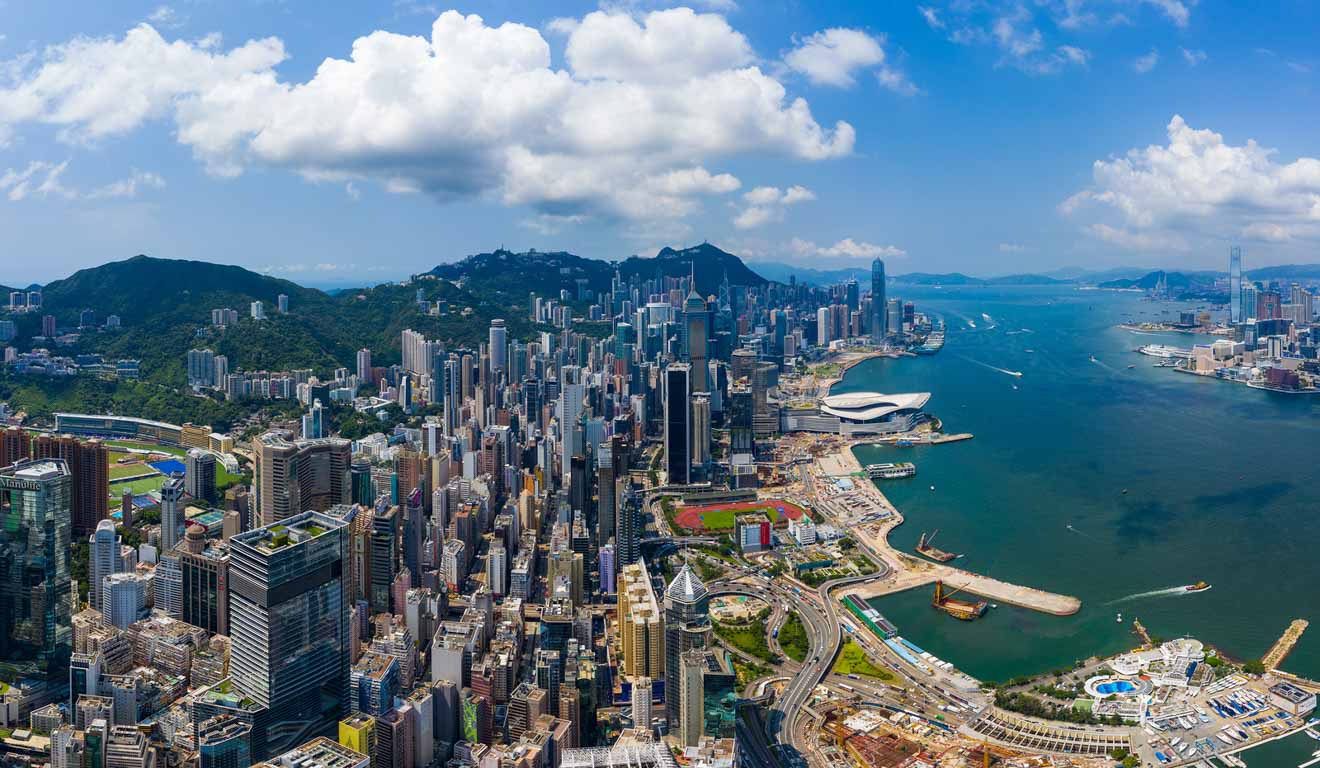 An aerial view of Hong Kong's skyscrapers and architectural landscape, with the harbor in the distance and a clear blue sky above.