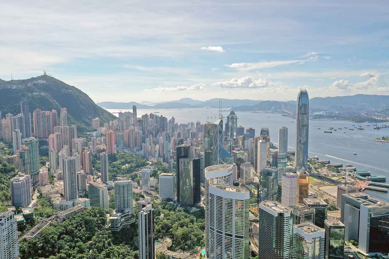 A clear day view of Hong Kong's expansive cityscape, featuring a mix of green hills and dense, high-rise buildings, with the harbor visible in the background.