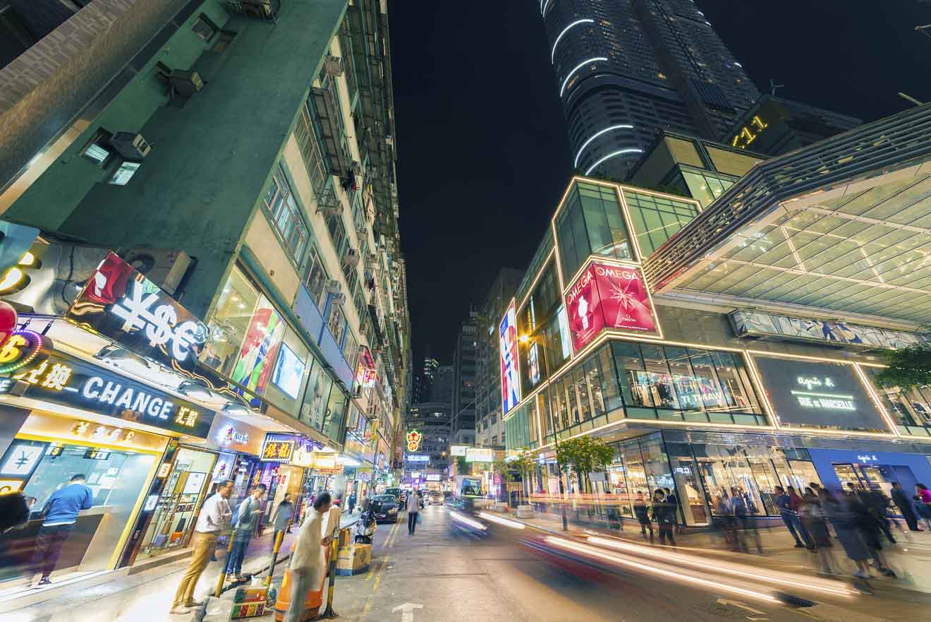 A bustling Hong Kong street at night, with neon signs illuminating the scene and people walking past the various shops and businesses.