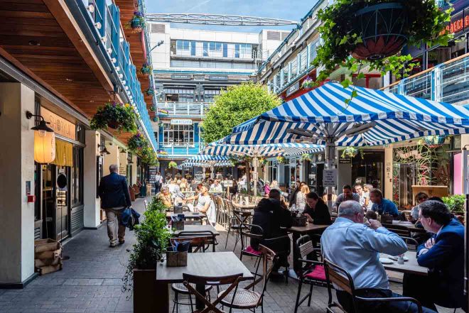 A lively outdoor dining area with striped umbrellas, people enjoying meals at various restaurants, and a clear blue sky above