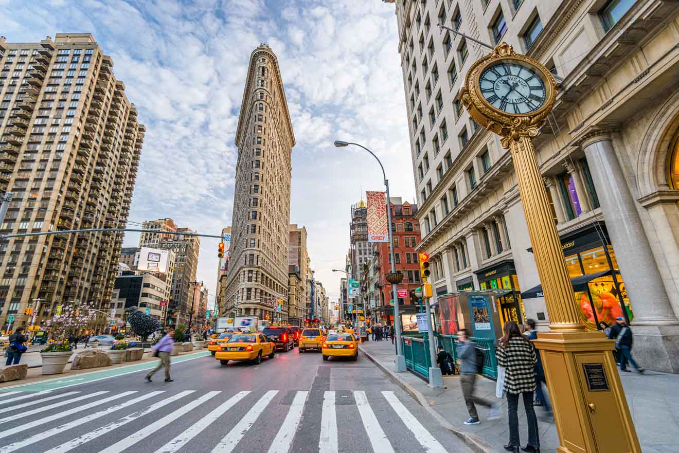 The Flatiron Building in New York City with a clock post in the foreground, bustling street, and yellow taxis.