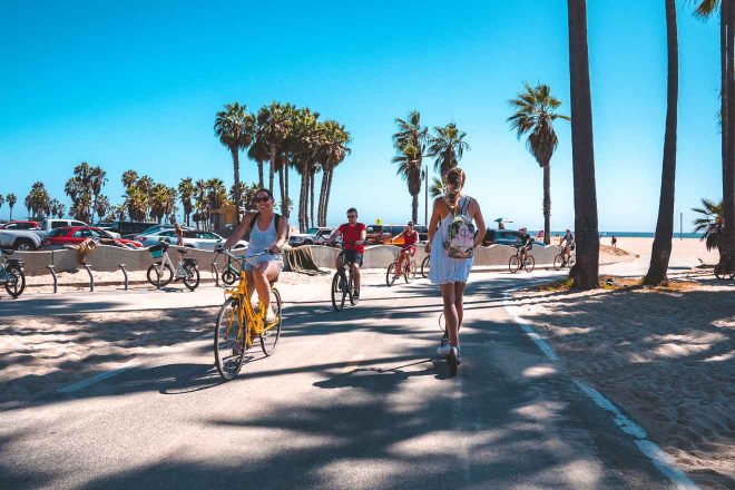 Casual beachside ambiance with people biking and walking along a path lined with palm trees on a bright, sunny day in Venice Beach, Los Angeles.

