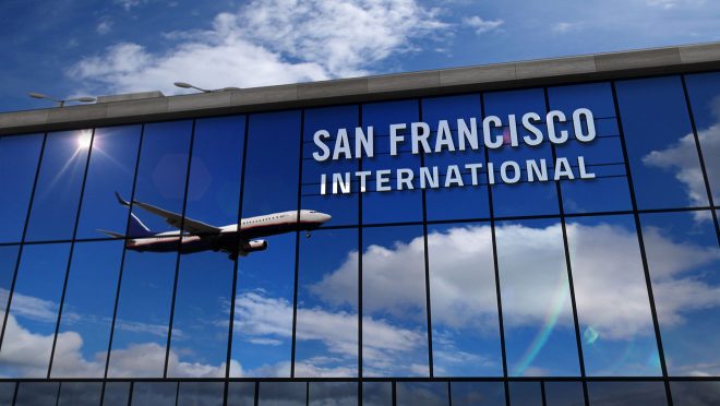 An airplane in flight captured against the backdrop of a partly cloudy sky, with the bold lettering of 'San Francisco International' reflecting on the facade of the airport terminal building.