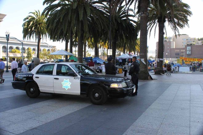 A San Francisco police car parked on a plaza with palm trees, with people and market stalls in the background
