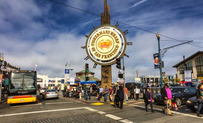 Bustling street scene at Fisherman's Wharf in San Francisco with pedestrians, cyclists, and vehicles under the iconic Fisherman's Wharf sign on a partly cloudy day