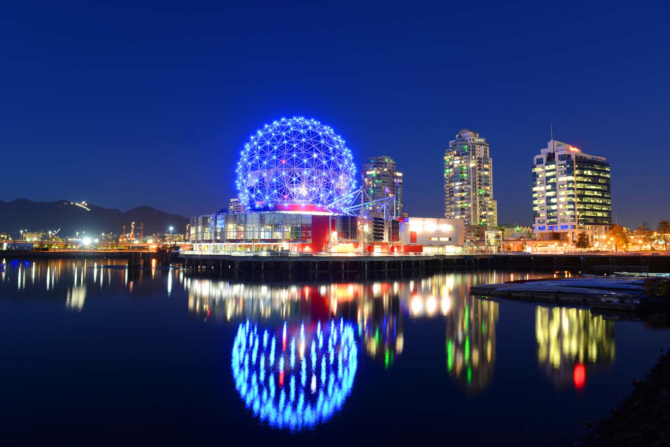 Nighttime cityscape of Vancouver featuring the Science World geodesic dome lit in vibrant blue with its reflection in the calm water.