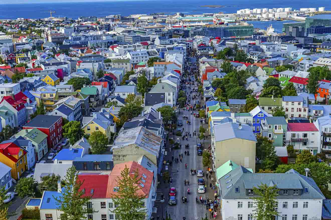 Aerial view of a bustling street in Reykjavik, Iceland, lined with colorful houses and a busy thoroughfare filled with pedestrians and vehicles, with the cityscape and ocean in the distance