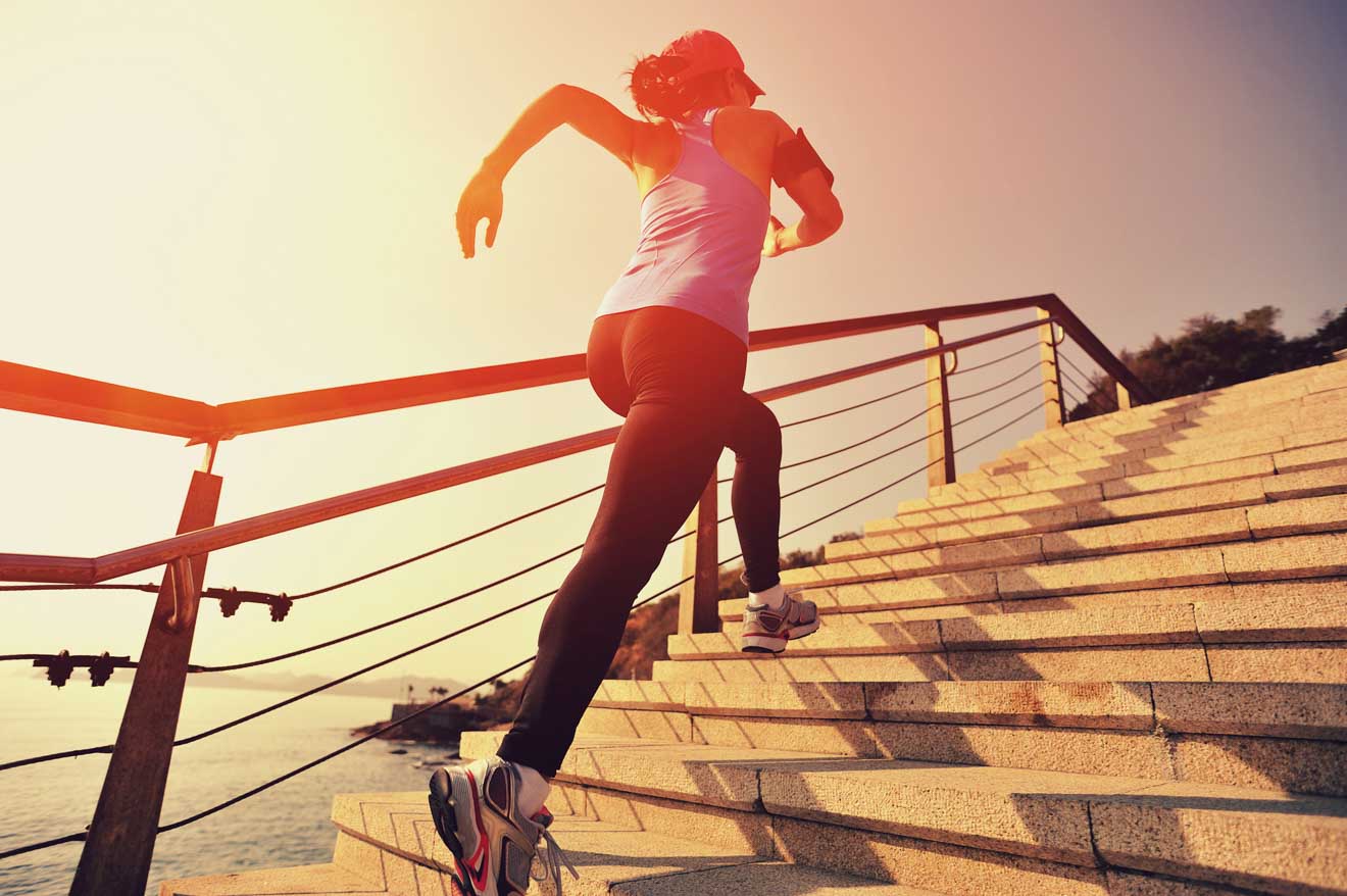 A person in athletic clothing runs up a staircase outdoors near a body of water, with the sun low in the sky.