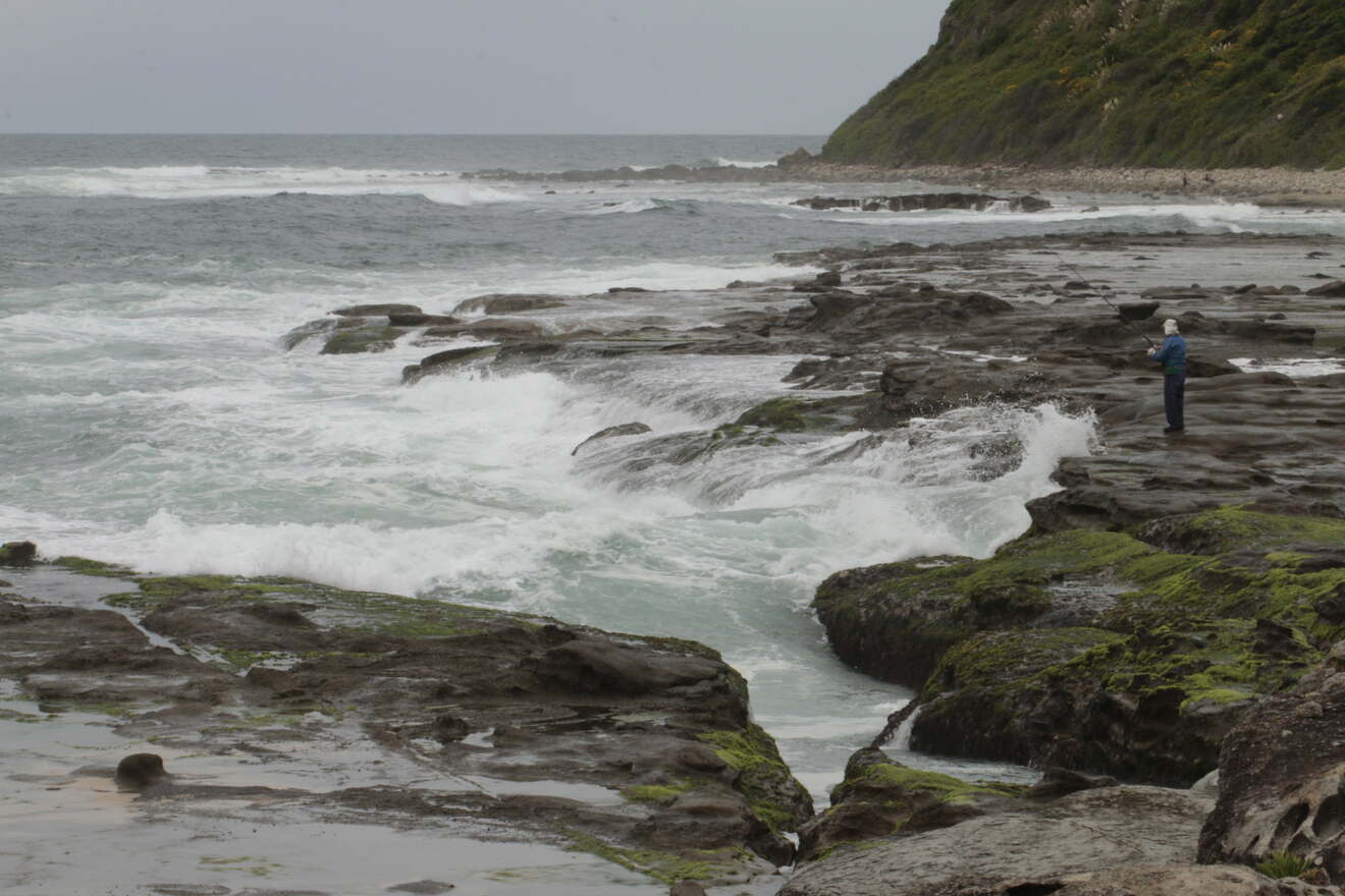 A person standing on a rocky, wave-swept coastline under a cloudy sky. Waves crash against the shore, with green-topped rocks visible in the foreground.