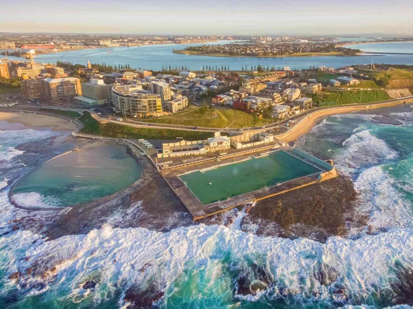 A coastal cityscape with a beach, ocean waves, and a distinctive rectangular ocean pool surrounded by residential buildings and a distant harbor view.