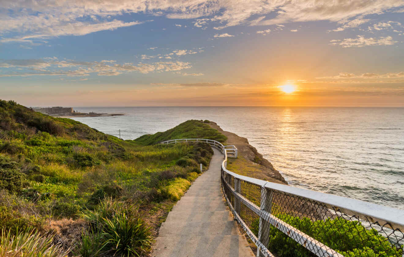 A narrow path with a railing leads down a hill towards the ocean at sunset, surrounded by green vegetation under a partly cloudy sky.