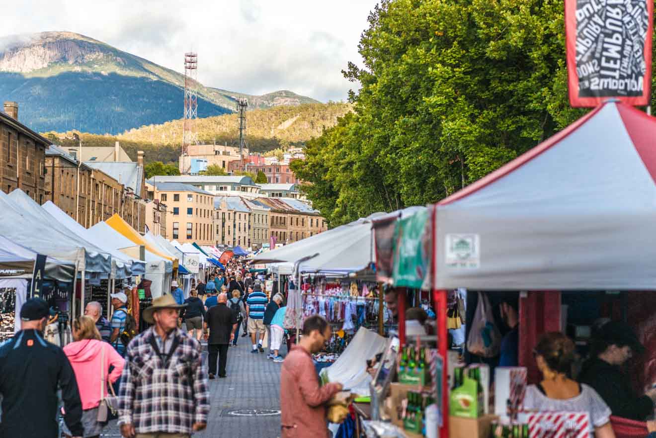 Outdoor market scene with people walking among stalls offering goods under colorful canopies. A mountain and buildings are visible in the background.