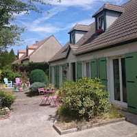 A charming two-story house with green shutters, dormer windows, and a front garden with potted plants on a sunny day. Outdoor furniture is visible in the background.