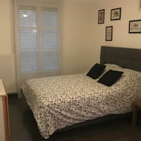 A bedroom with a double bed, two black pillows, a patterned bedspread, a window with blinds, and framed pictures on the wall.