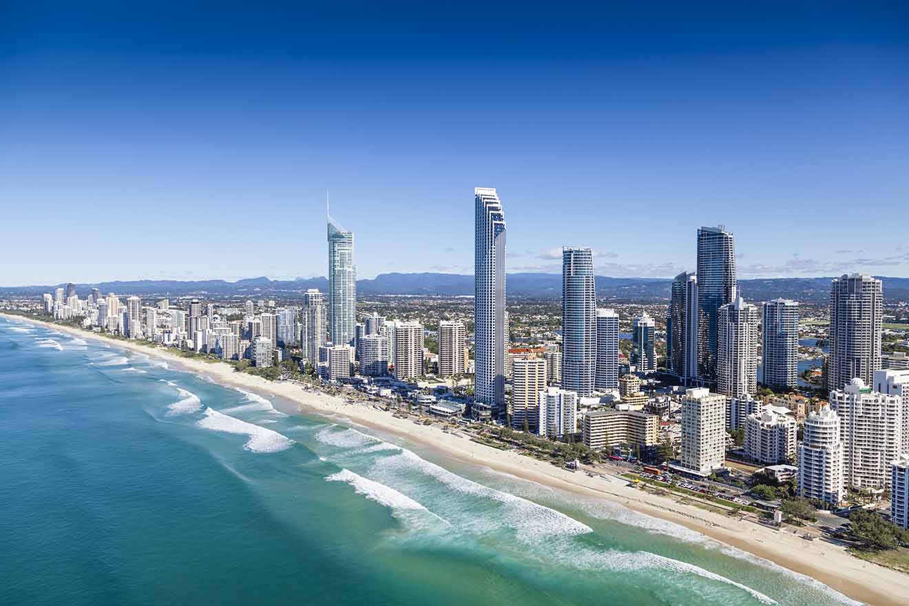 Aerial view of a coastal cityscape with tall modern buildings alongside a sandy beach and ocean waves under a clear blue sky.