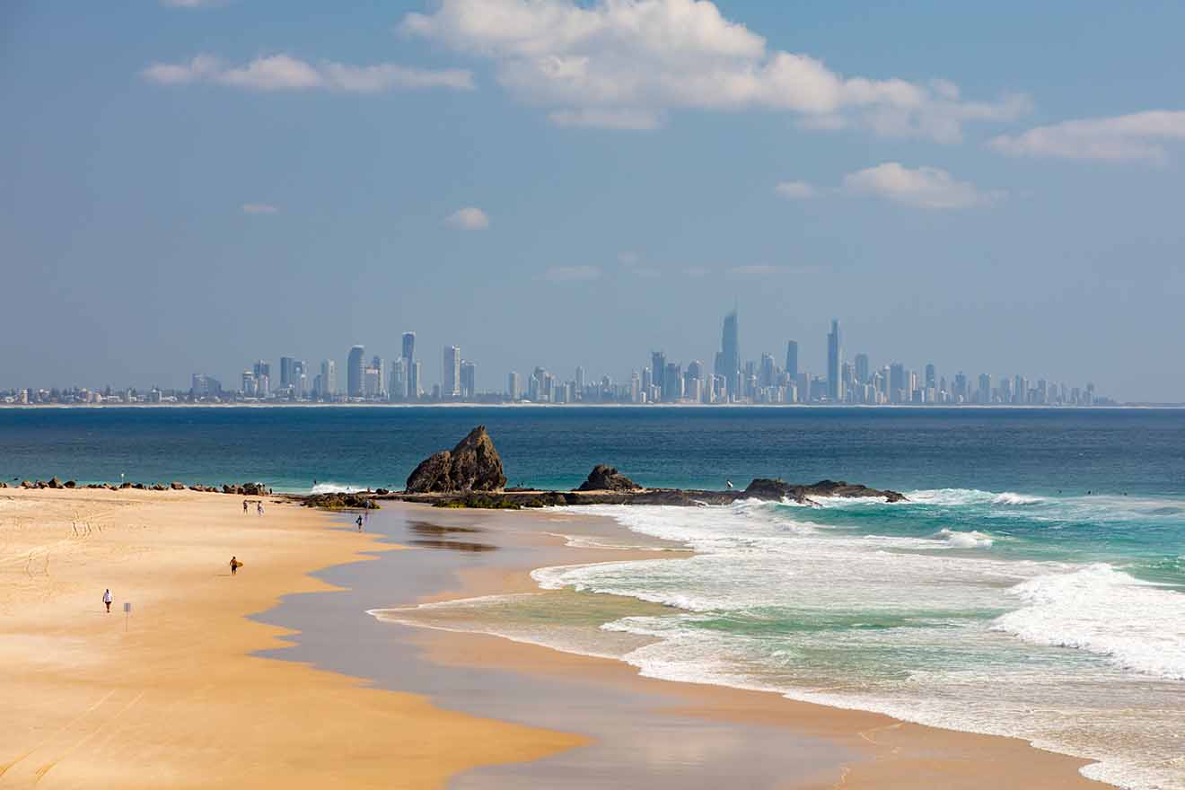 A sandy beach with scattered people in the foreground, waves crashing onshore. The background shows a distant city skyline under a clear blue sky with a few clouds.