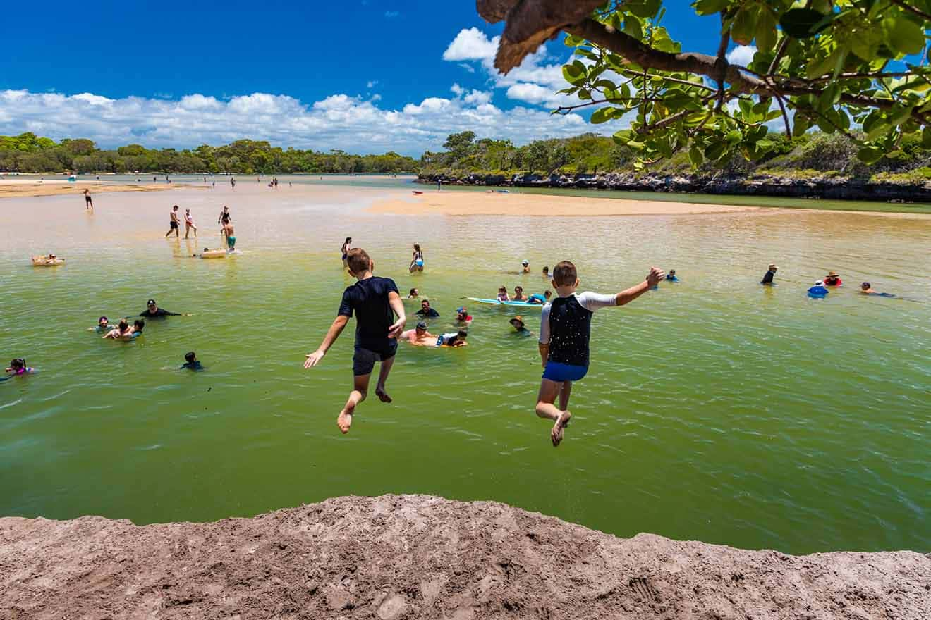 Children jumping into a green lagoon while others swim and play in the water under a blue sky with scattered clouds. Trees line the background.