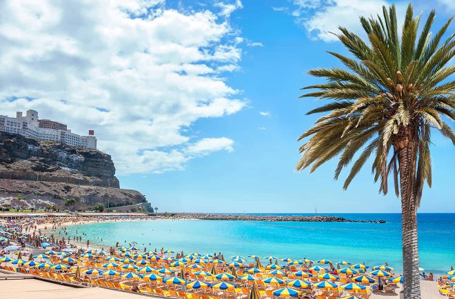 A beach with numerous yellow and blue umbrellas, clear blue water, a palm tree in the foreground, and cliffs with buildings in the background under a partly cloudy sky.