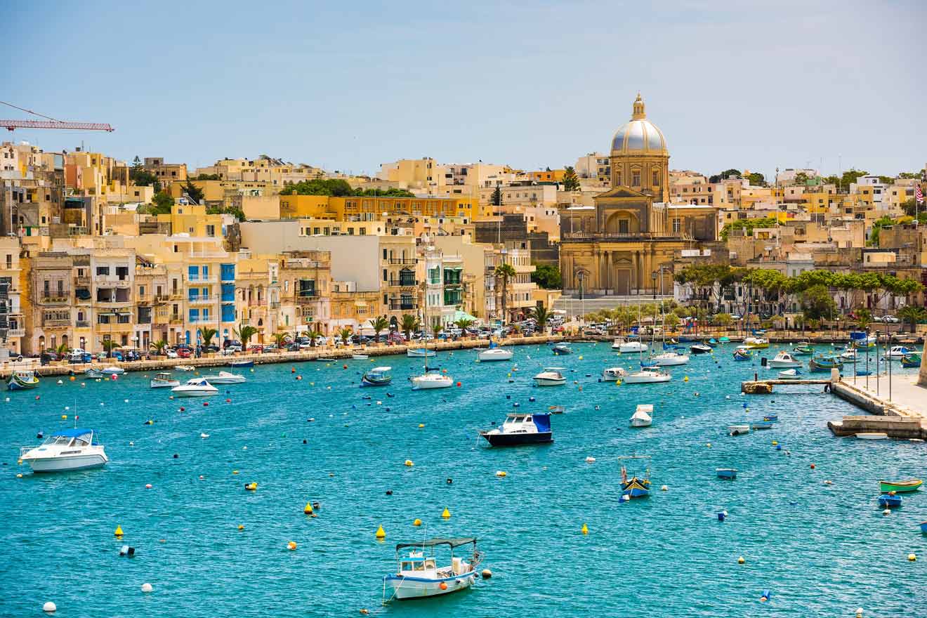 A scenic view of a harbor with small boats on turquoise water and colorful buildings in the background, featuring a prominent domed church.