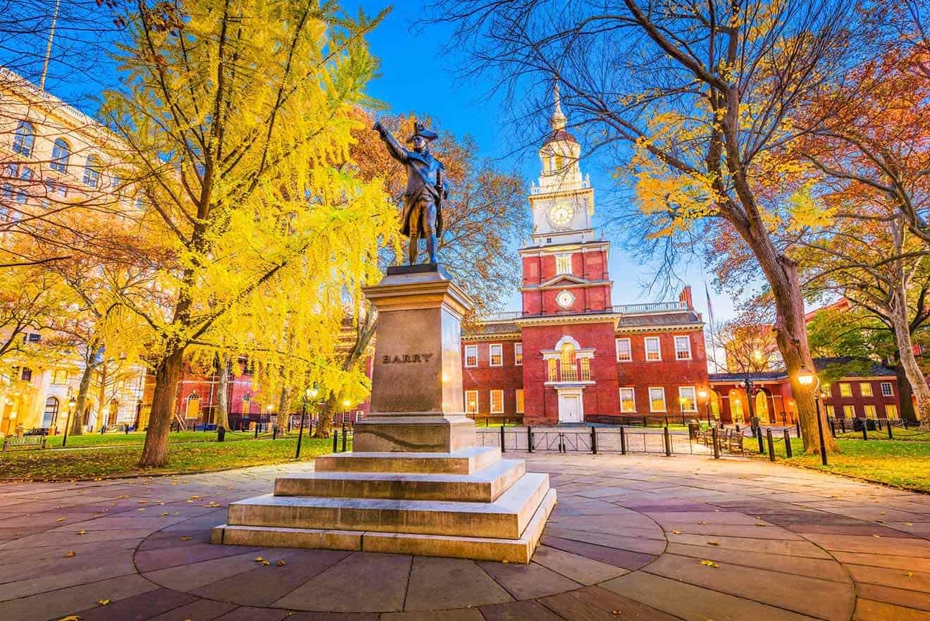 The historic Independence Hall in Philadelphia with the Commodore Barry statue in the foreground surrounded by autumn foliage under twilight.