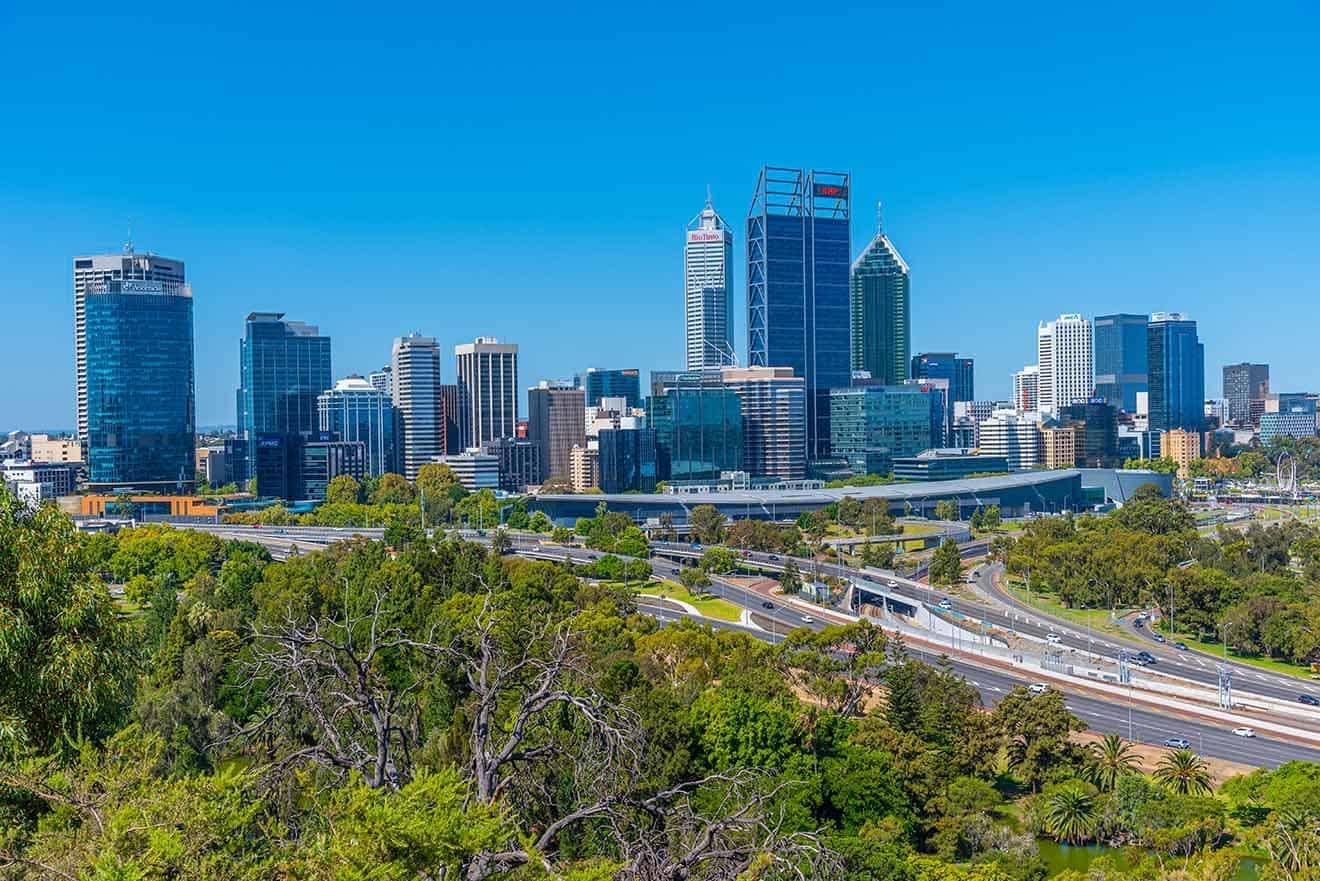 A daytime cityscape with modern high-rise buildings, clear blue skies, and lush greenery in the foreground