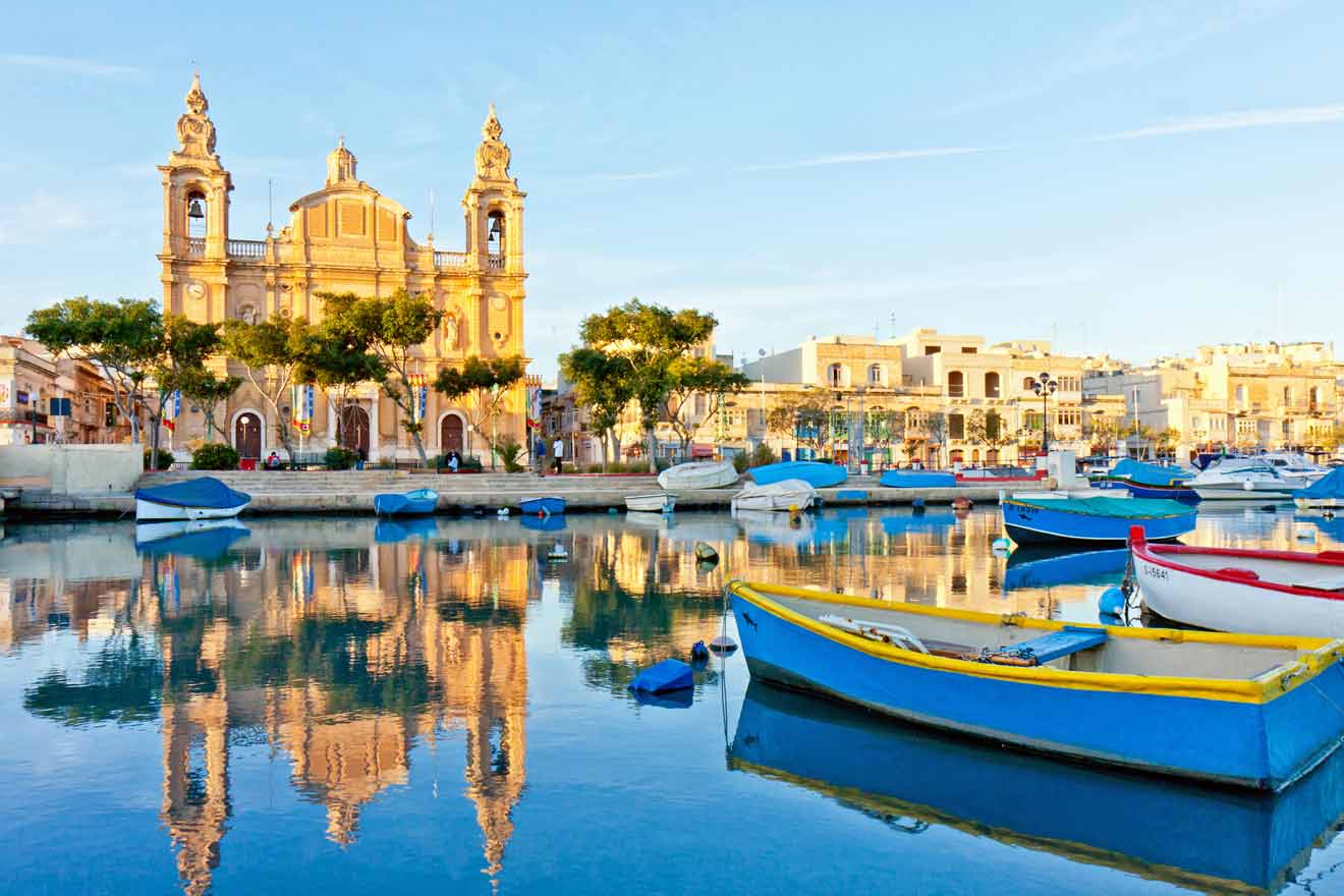 A waterfront scene with colorful boats on a calm waterway reflecting a historic church and surrounding buildings under a clear blue sky.