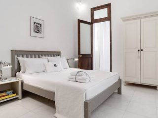 A neatly arranged bedroom with a double bed, bedside tables with lamps and books, an open wooden door, and a closed wardrobe. White walls and bedding create a clean, minimalist look.